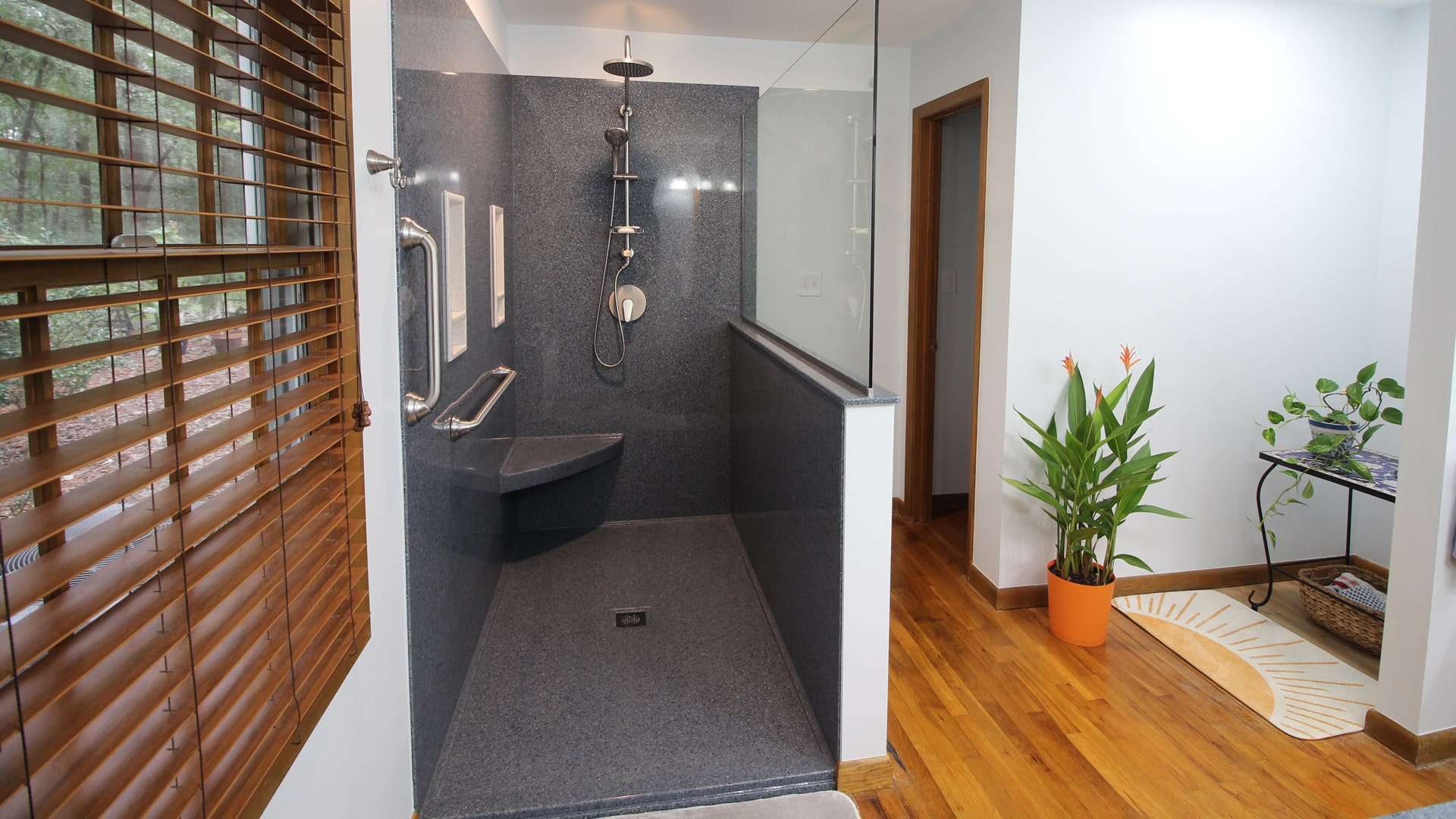 Modern bathroom featuring a dark walk-in shower, built-in bench, rainfall showerhead, grab bars, wood floor, plant, table, and large window.