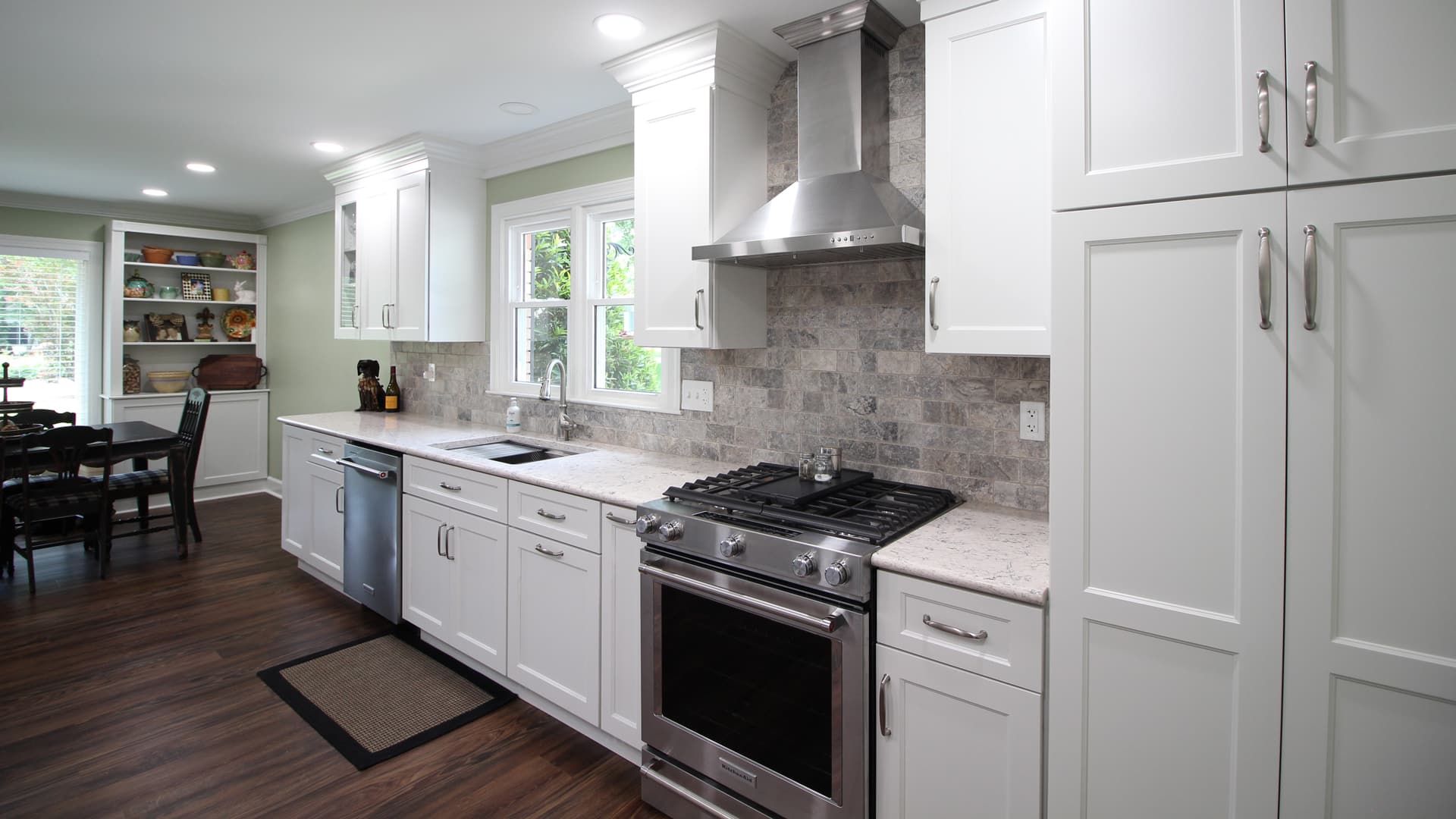 Modern kitchen with white cabinets, stainless appliances, gas stove, marble countertops, dark wood floors. Dining area behind.