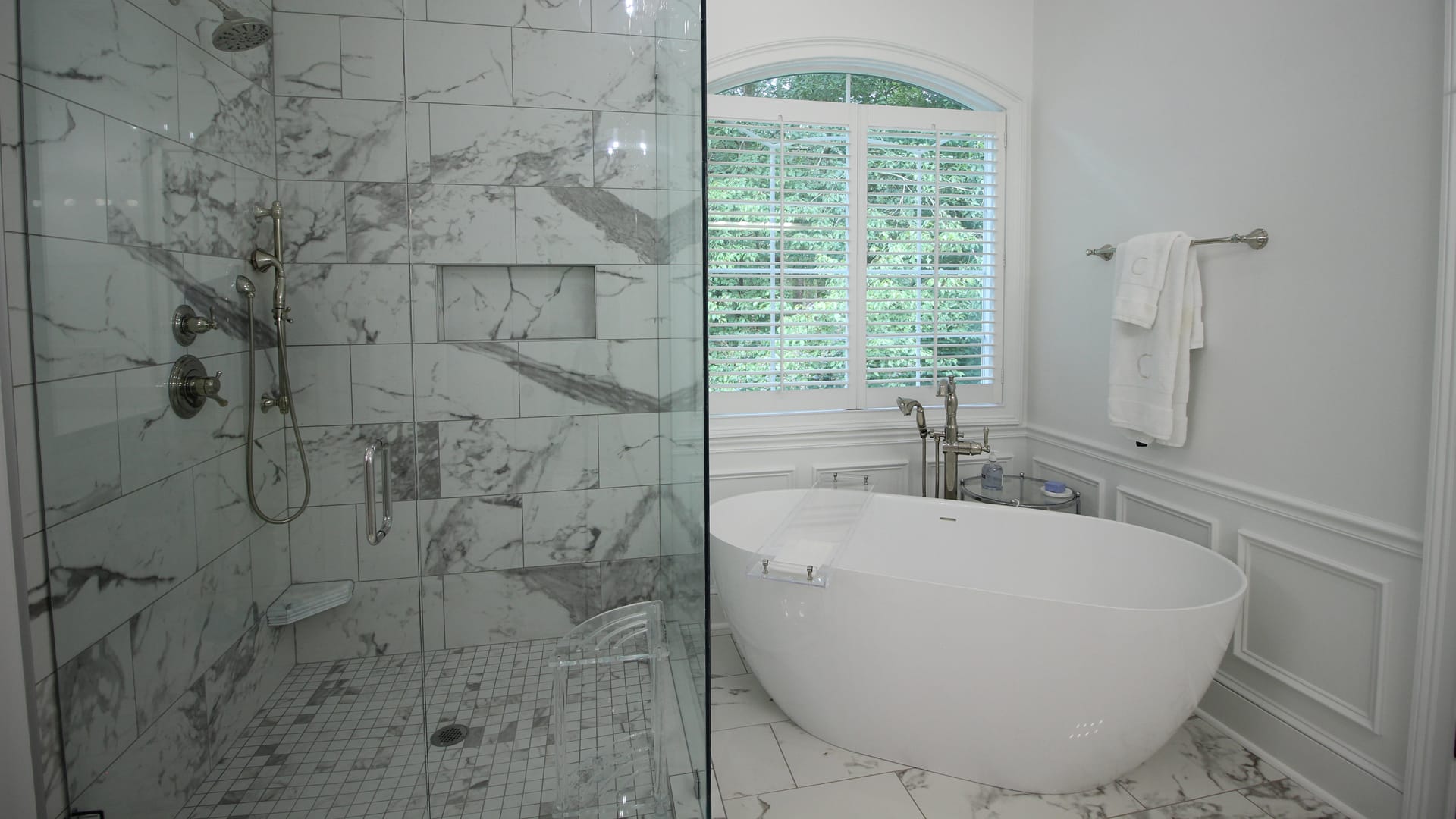 Modern bathroom featuring a glass-enclosed marble shower, freestanding tub, window with white shutters, tiled floor, and silver towel rack.
