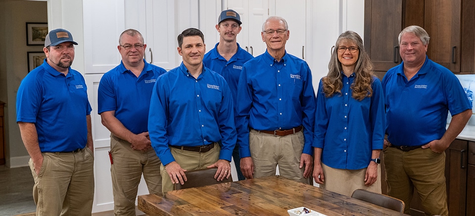 Seven people in matching blue shirts and khaki pants smile together in a kitchen with white and wood cabinets, near a wooden table.