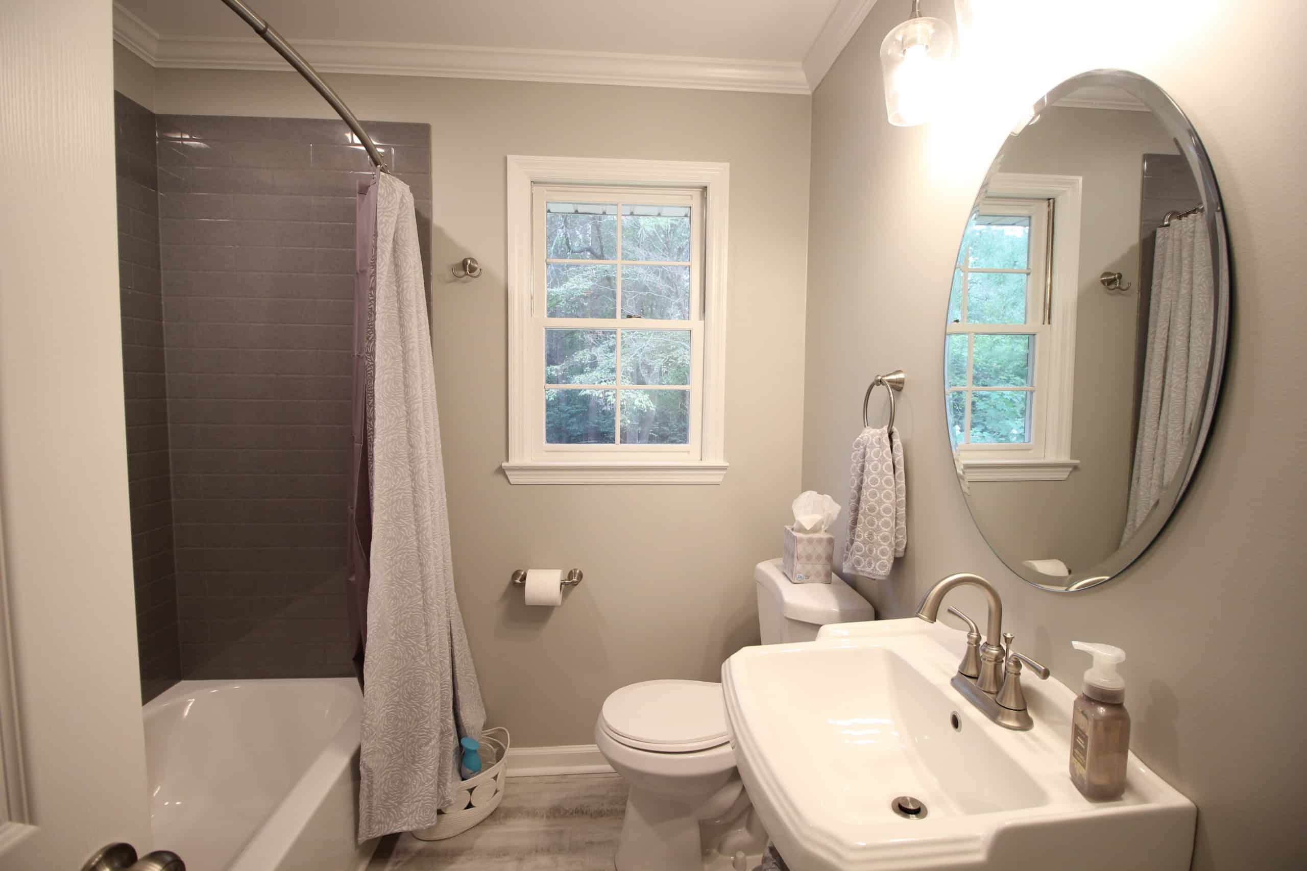 Modern bathroom with gray tiled shower, white curtain, toilet, sink with silver faucet, round mirror, window, towel and soap visible.