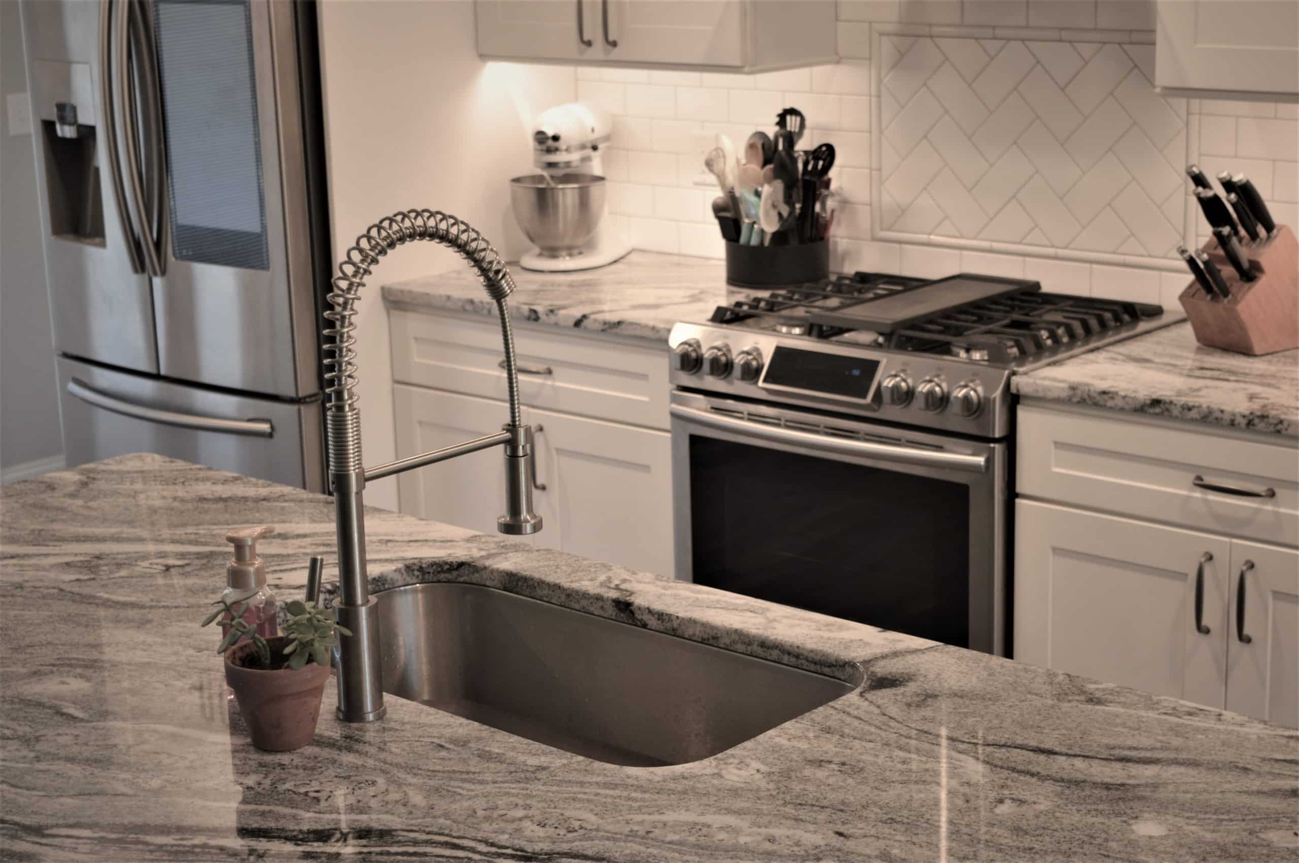 Modern kitchen with marble countertop, built-in sink, gooseneck faucet, white cabinets, stainless steel appliances, and a stand mixer.