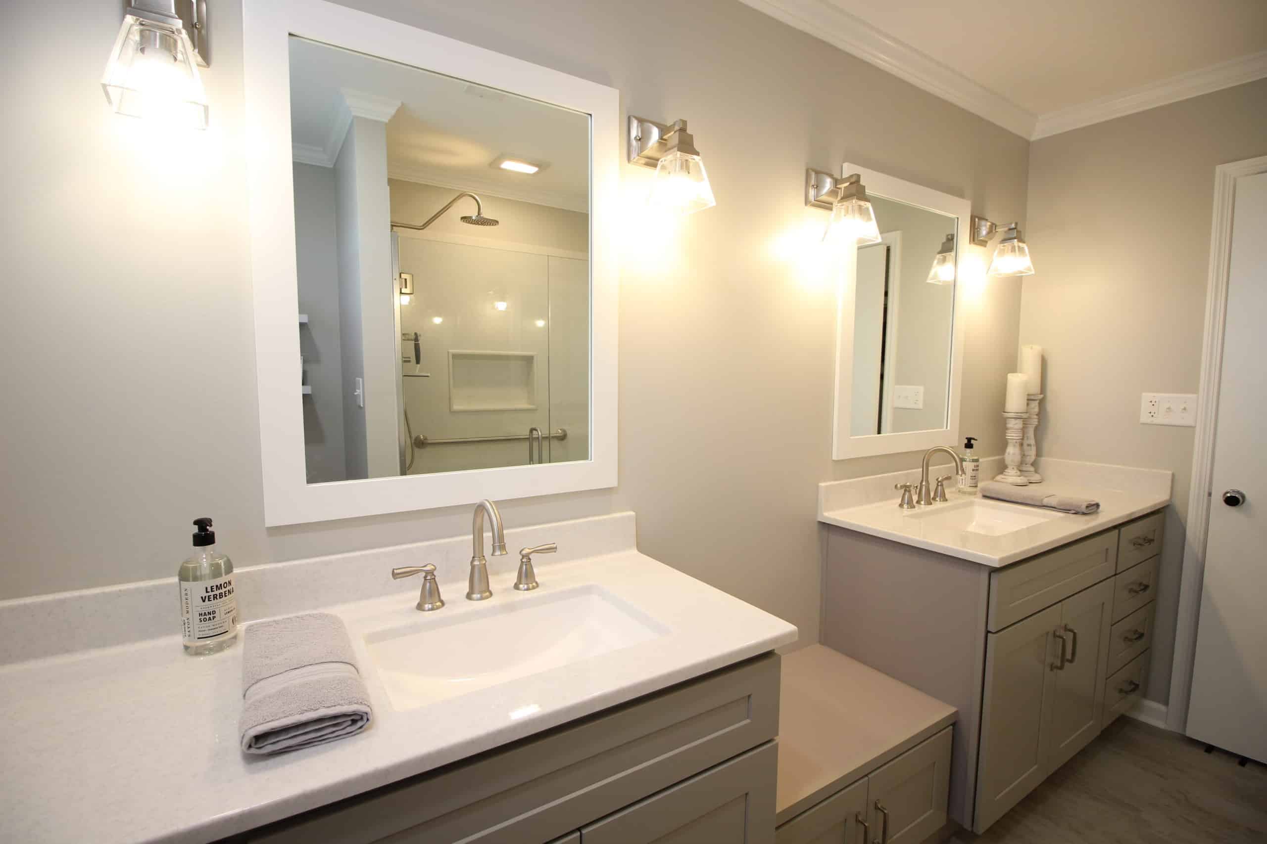 Modern bathroom with dual sinks, large mirrors, wall lights, gray cabinets, white countertop; shower visible in background.