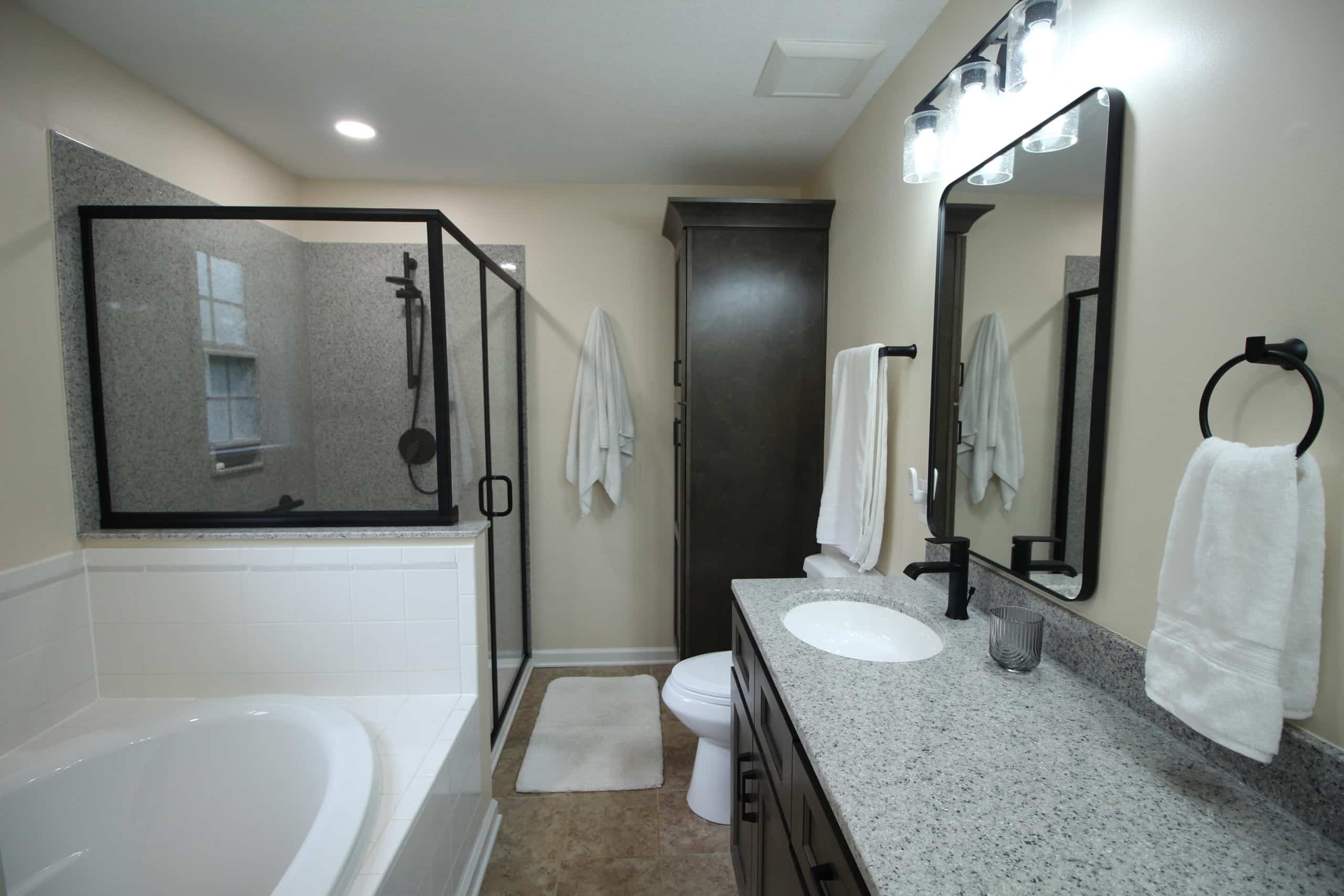 Modern bathroom featuring a glass shower, granite countertop, dark wood cabinets, white tub, towels on racks, and lit mirror above sink.