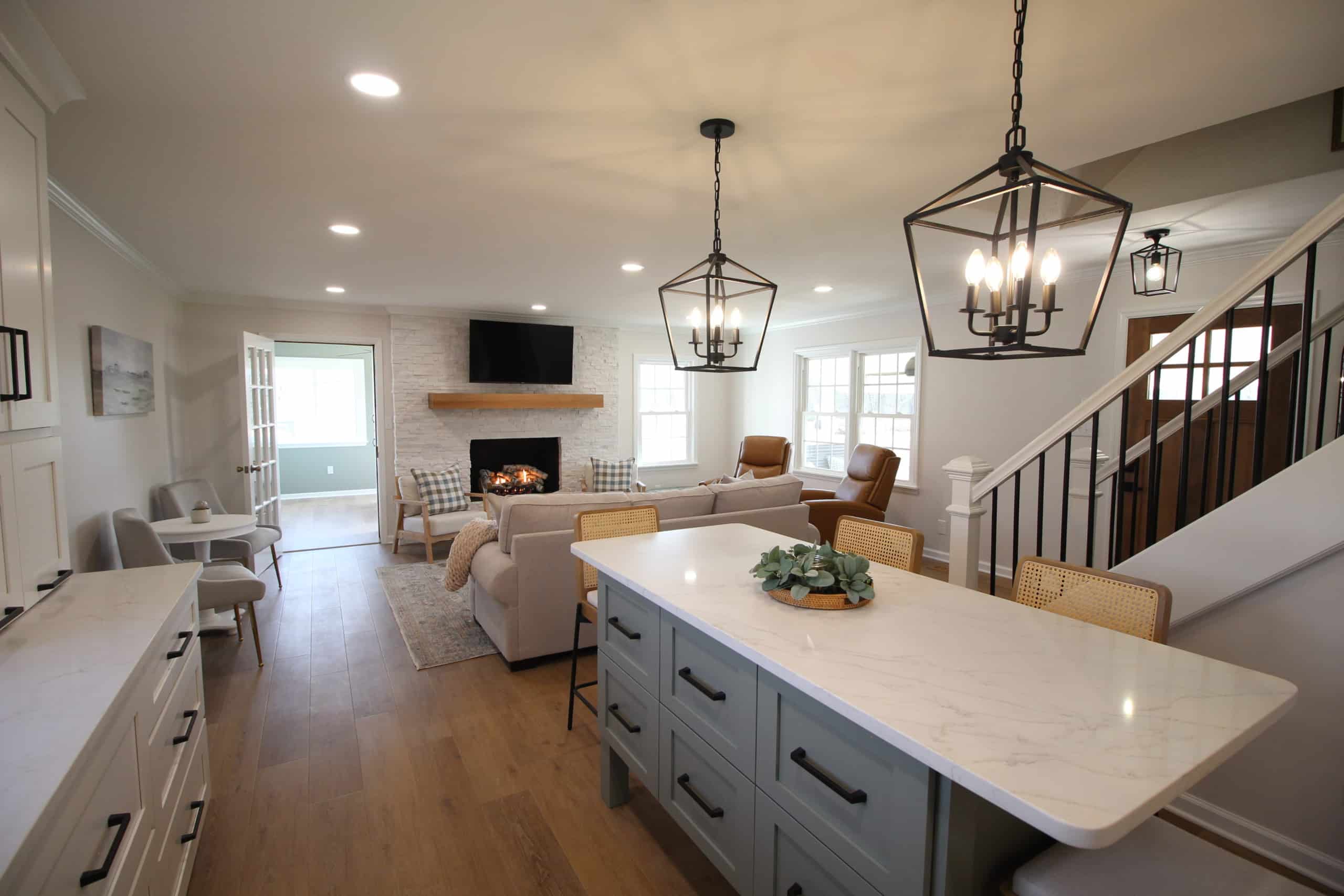 Modern open-concept kitchen and living room with a white island, pendant lights, light wood floors, fireplace, neutral furniture, large windows.