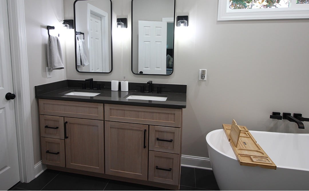 Modern bathroom with double sinks, black fixtures, rectangular mirrors, wall lights, towel rack, white tub with wood tray, white walls, black floor.