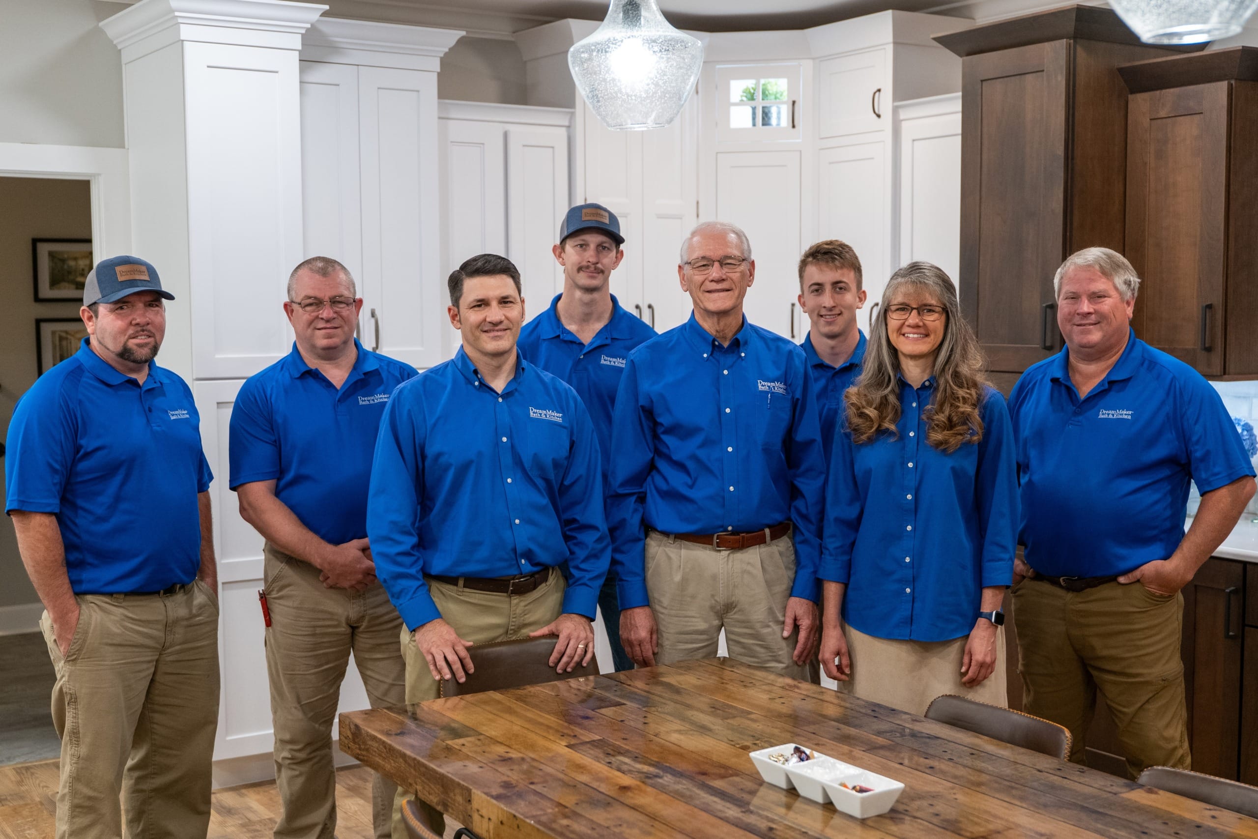 Eight people in matching blue shirts and khaki bottoms smile together in a bright kitchen; a wooden table with chairs is in front.