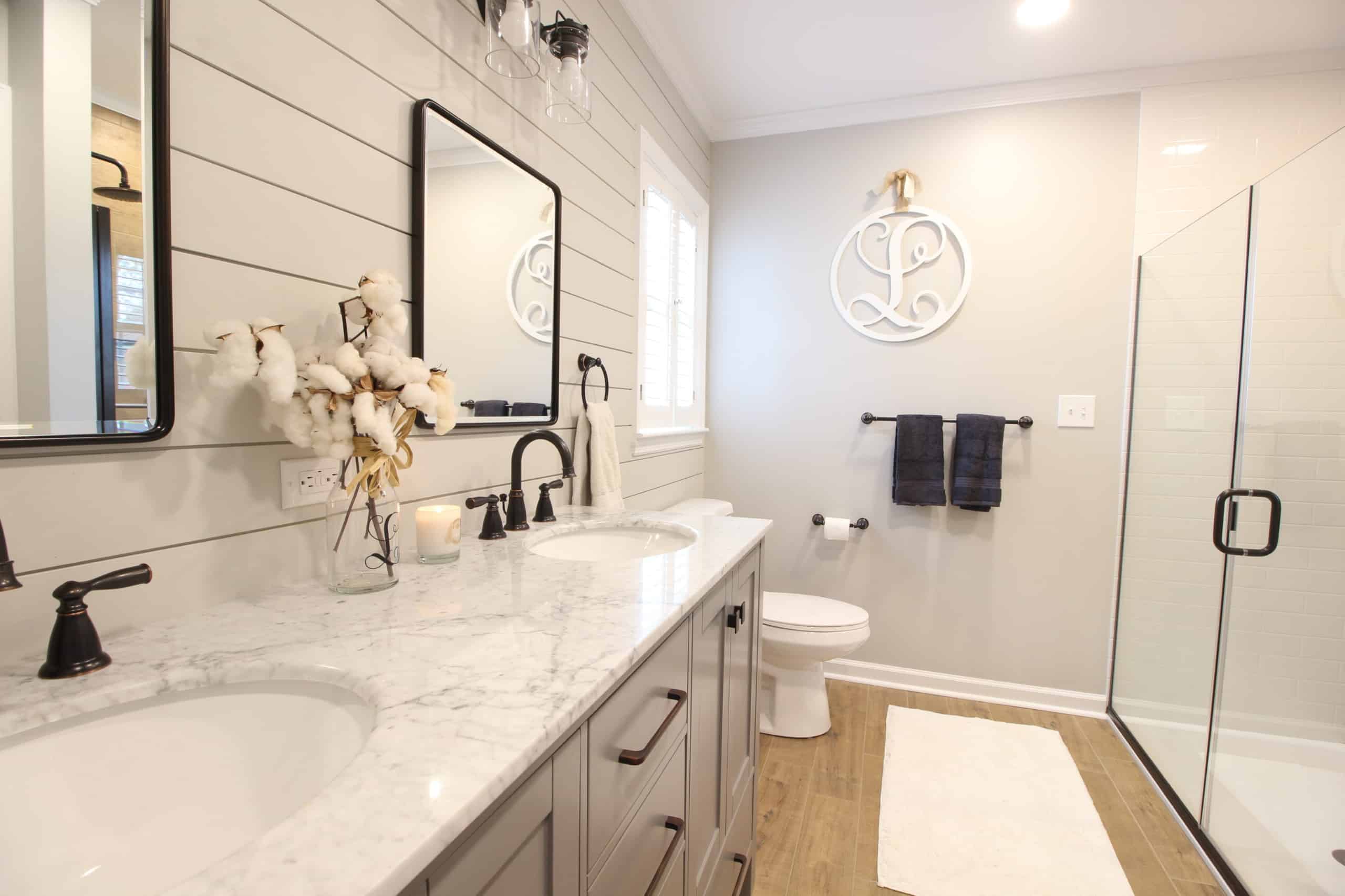 Modern bathroom with double sink vanity, marble countertop, black fixtures, round wall decor above toilet, glass shower, wood floor.