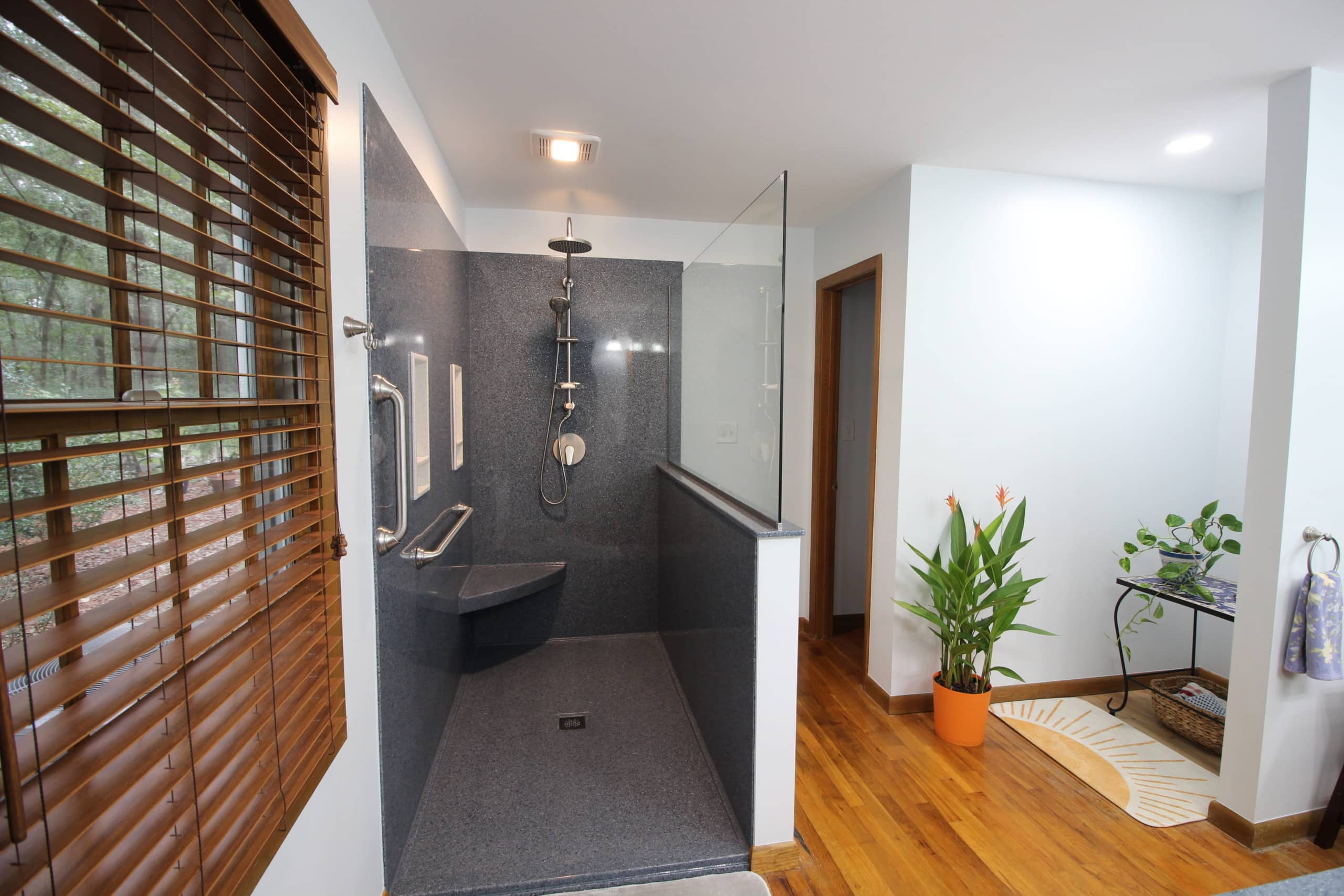 Modern bathroom with walk-in shower, gray walls, built-in seat, grab bars, handheld showerhead, wood floors, and window blinds.