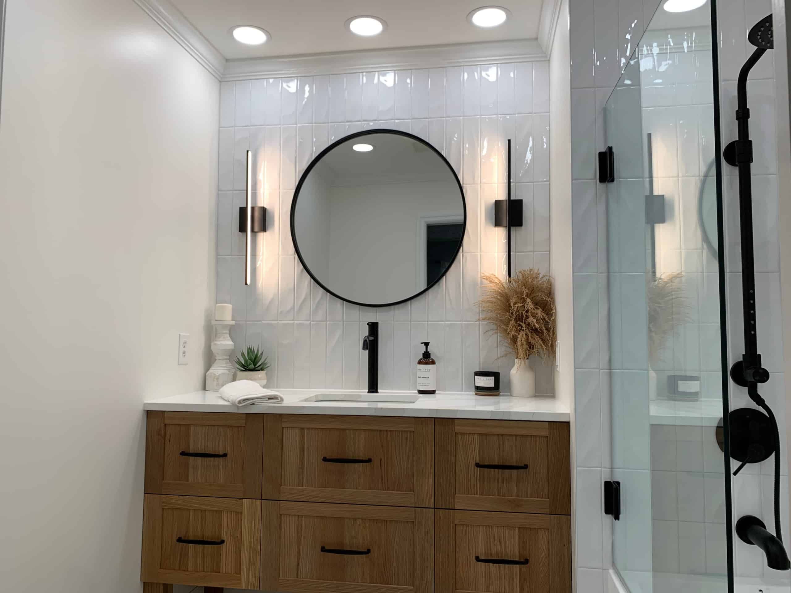 Modern bathroom with wooden vanity, round black-framed mirror, wall lights, white countertop, decor items, and partial glass shower.