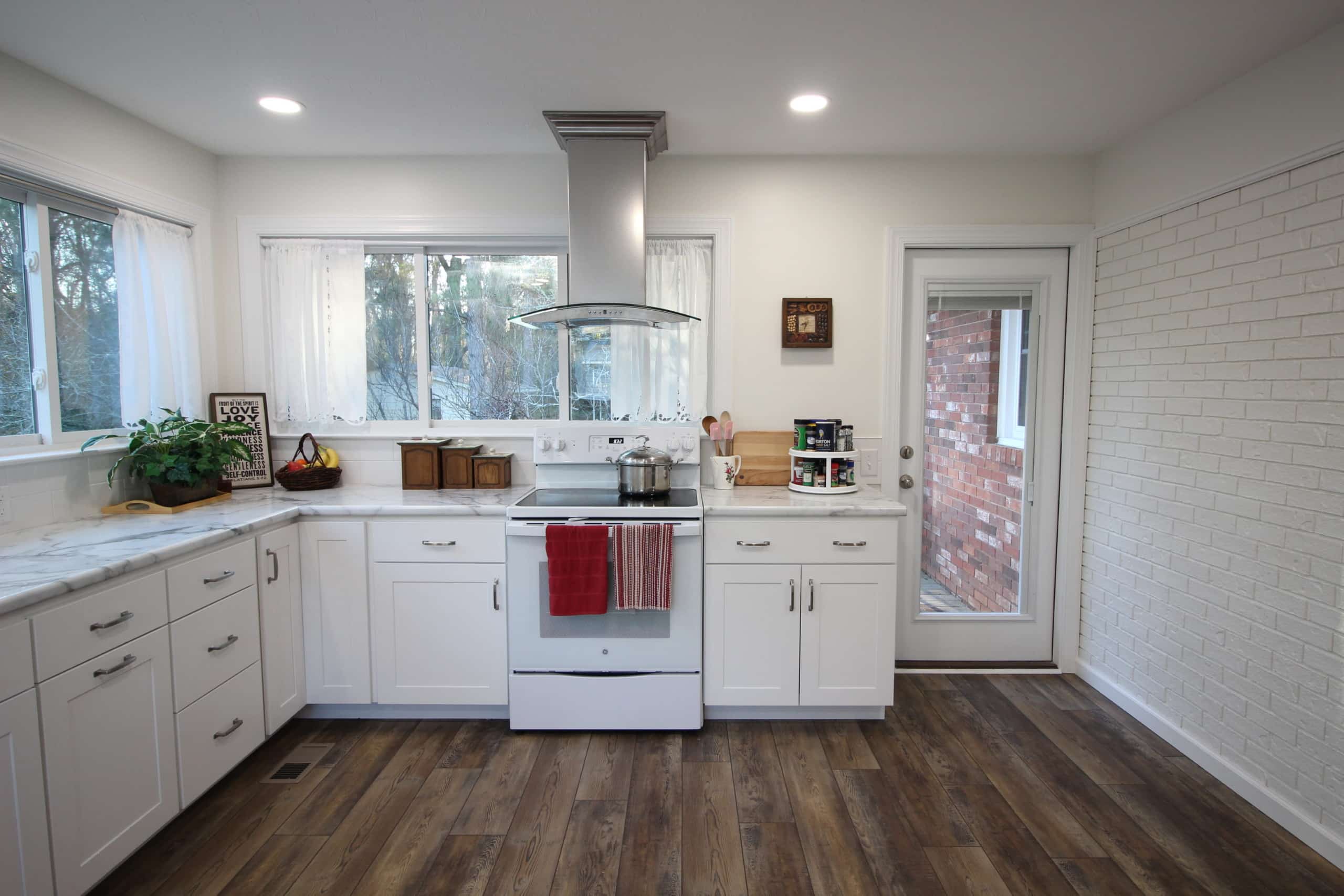 Bright modern kitchen with white cabinets, white stove, wooden floor, large windows, and door to outside; plants on counters.