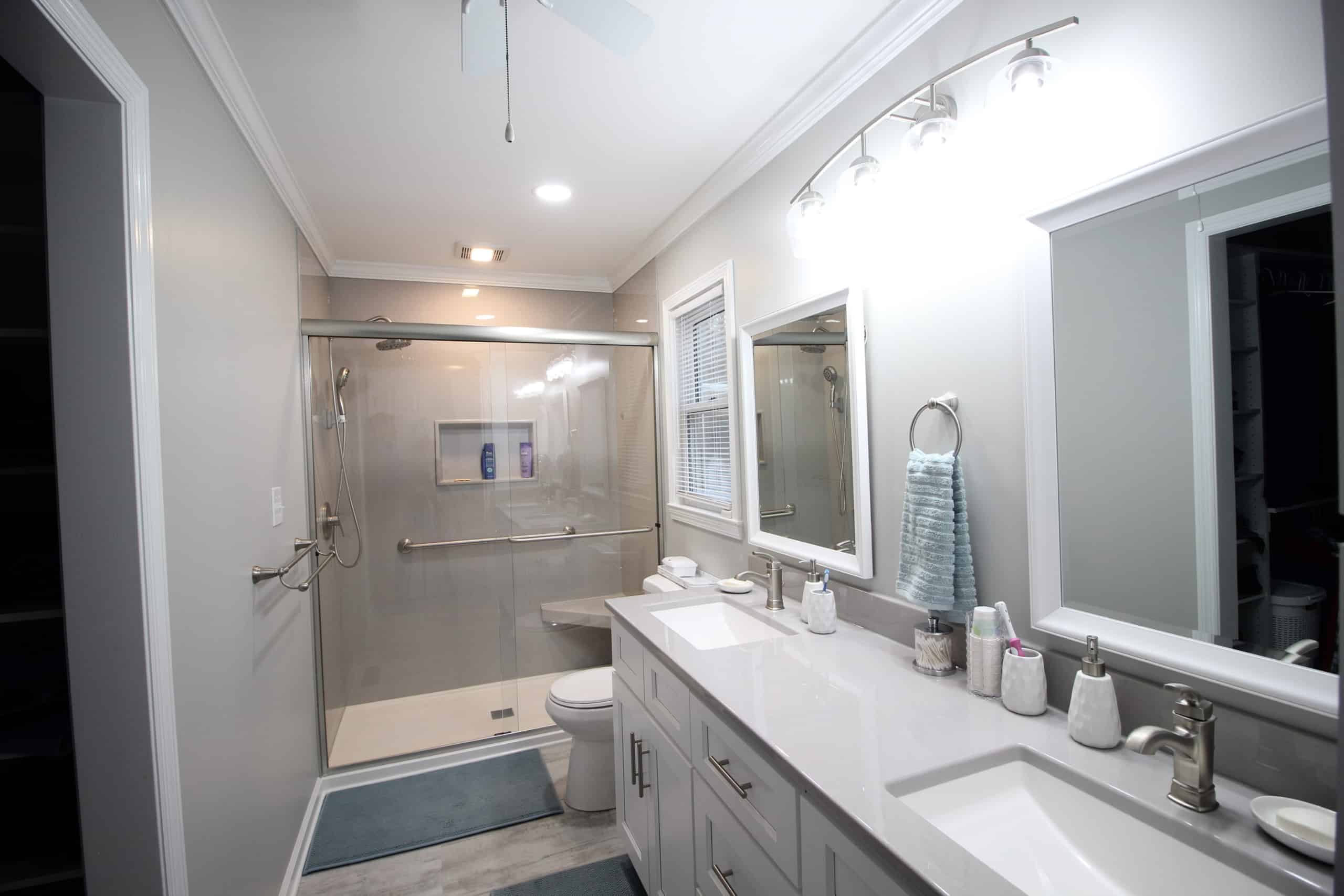 Modern bathroom with glass shower, white double-sink vanity, chrome fixtures, blue rugs, and bright lighting. Clean and tidy.