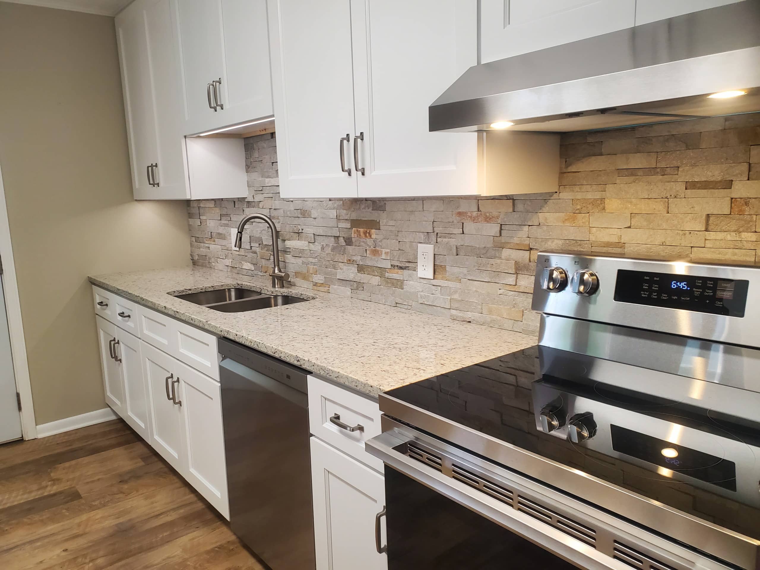 Modern kitchen featuring white cabinets, a stone backsplash, stainless steel appliances, double sink, and light-colored granite countertops. Wooden floor and under-cabinet lighting provide added brightness.