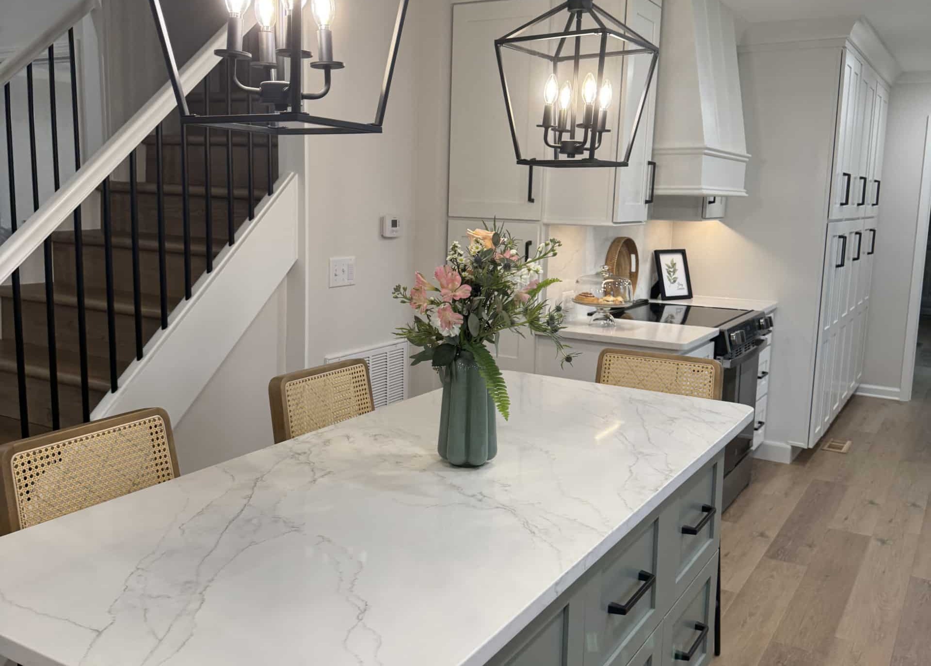 Modern kitchen with a marble island, two pendant lights, three chairs, a vase with flowers, stairs with black railing, wood flooring, and white cabinets.