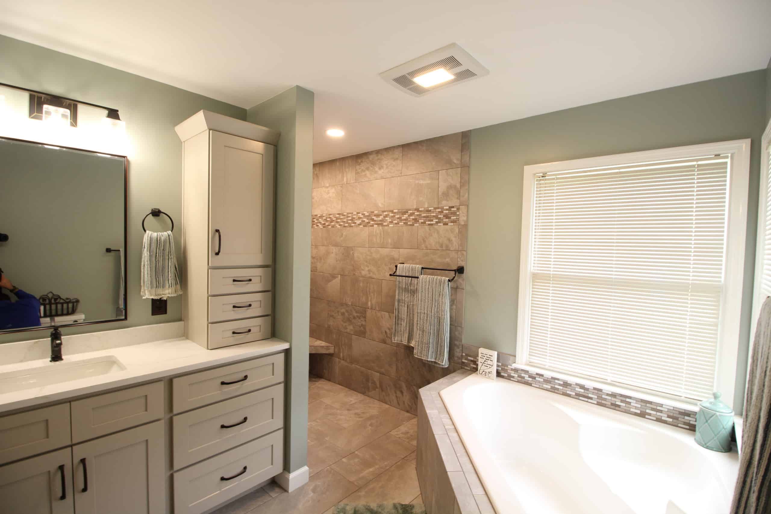 Modern bathroom with light gray cabinets, a large mirror, walk-in tiled shower featuring a built-in bench, white bathtub positioned by a window with blinds, and soft green walls. Towels are neatly hung beside the shower.