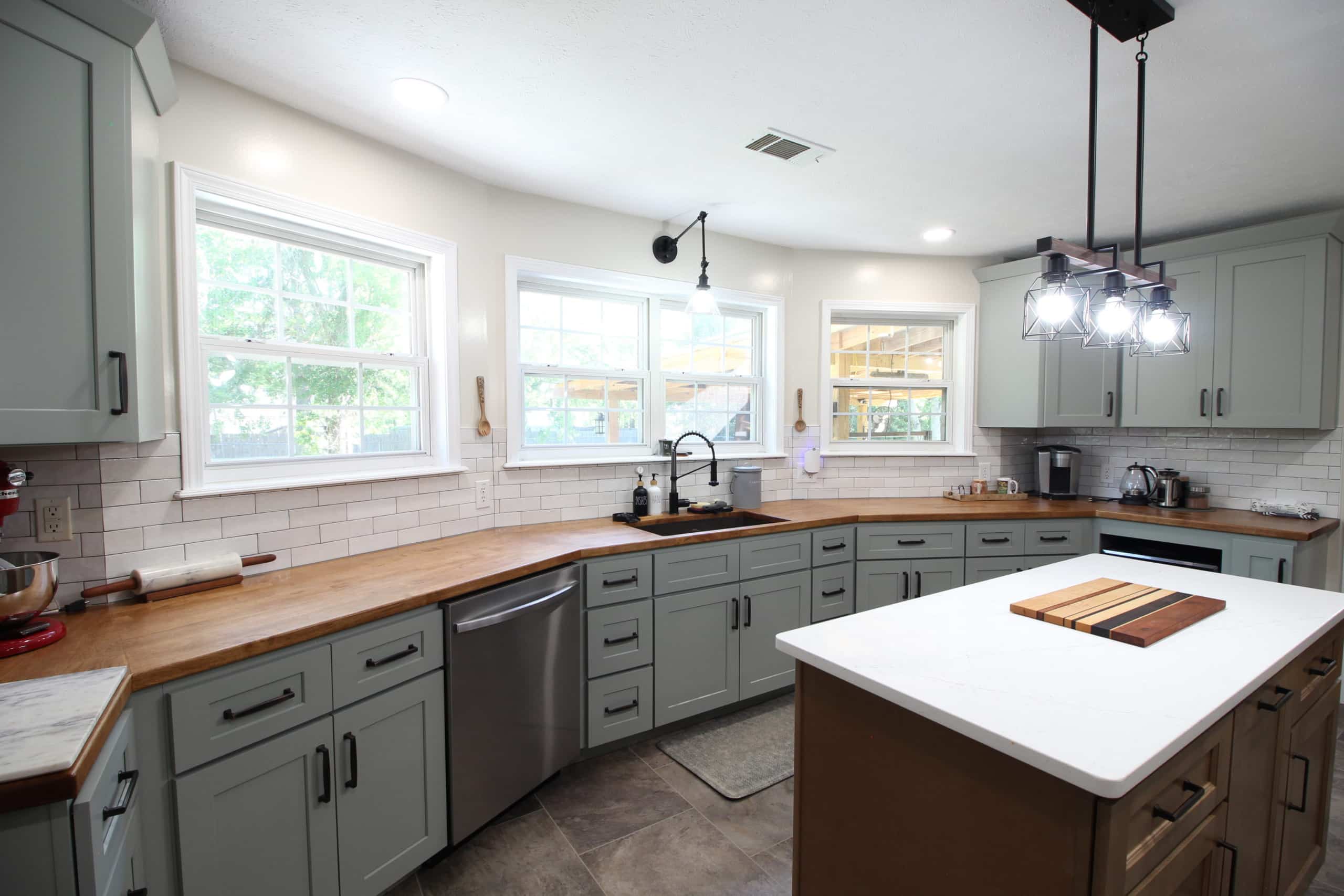 Modern kitchen featuring light green cabinets, wooden countertops, white subway tile backsplash, stainless steel appliances, a central island with a white surface, and large windows that allow abundant natural light.