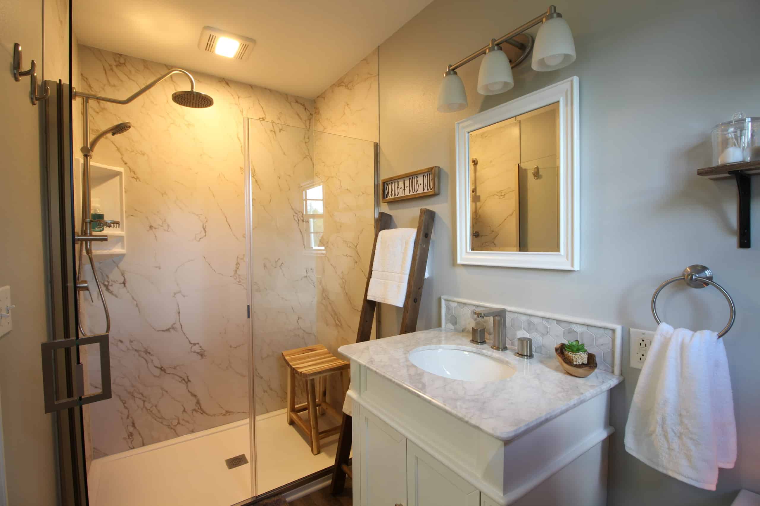 Modern bathroom featuring a marble shower with glass doors, a wooden bench, and a white vanity topped with marble. A mirror hangs above the vanity, next to a towel rack and shelves displaying decor and toiletries. The room is bright and neatly organized.