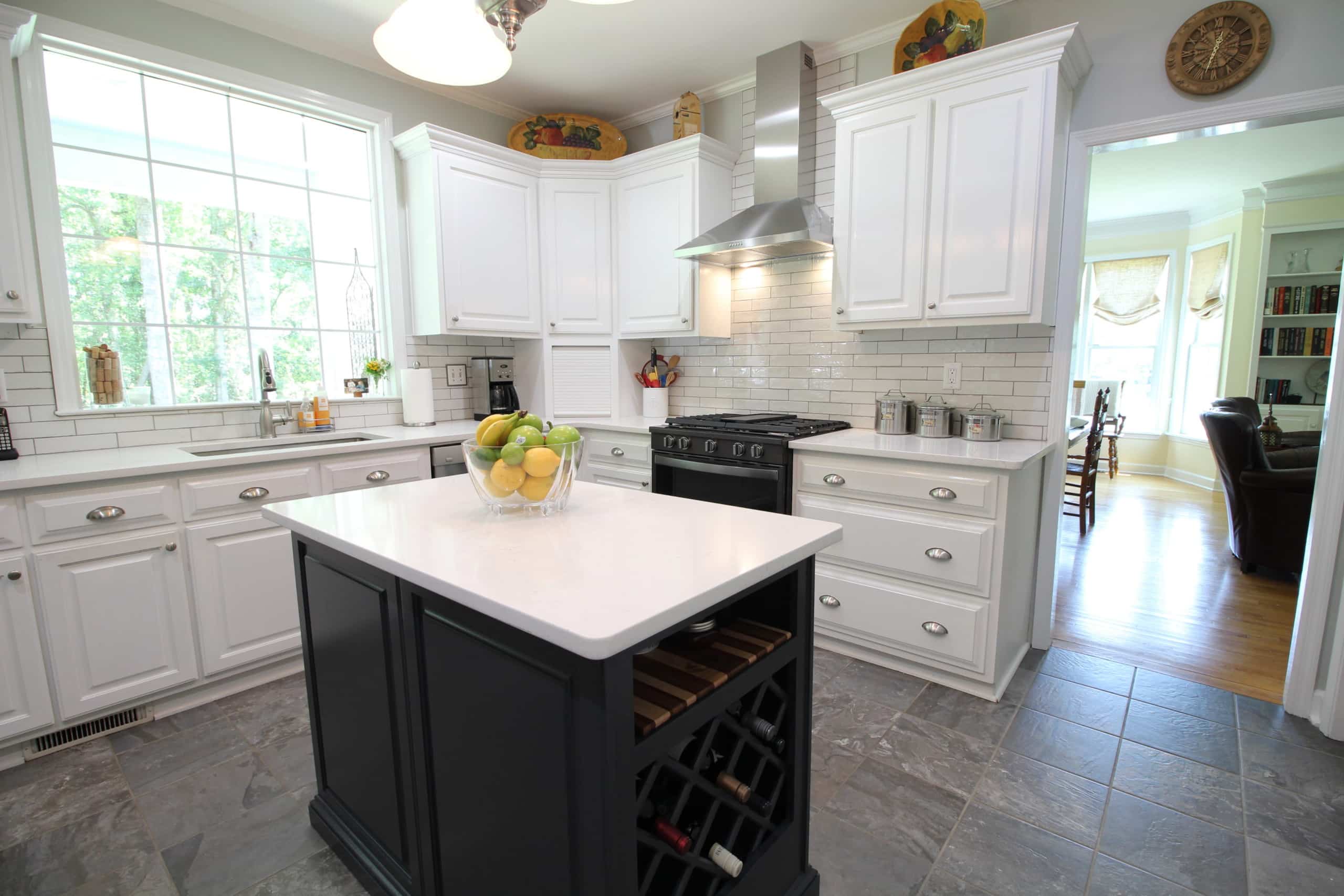 Bright kitchen featuring white cabinets, stainless steel appliances, and a black island with wine storage and a fruit bowl on top. A large window lets in natural light, and the dining and living areas can be seen in the background.