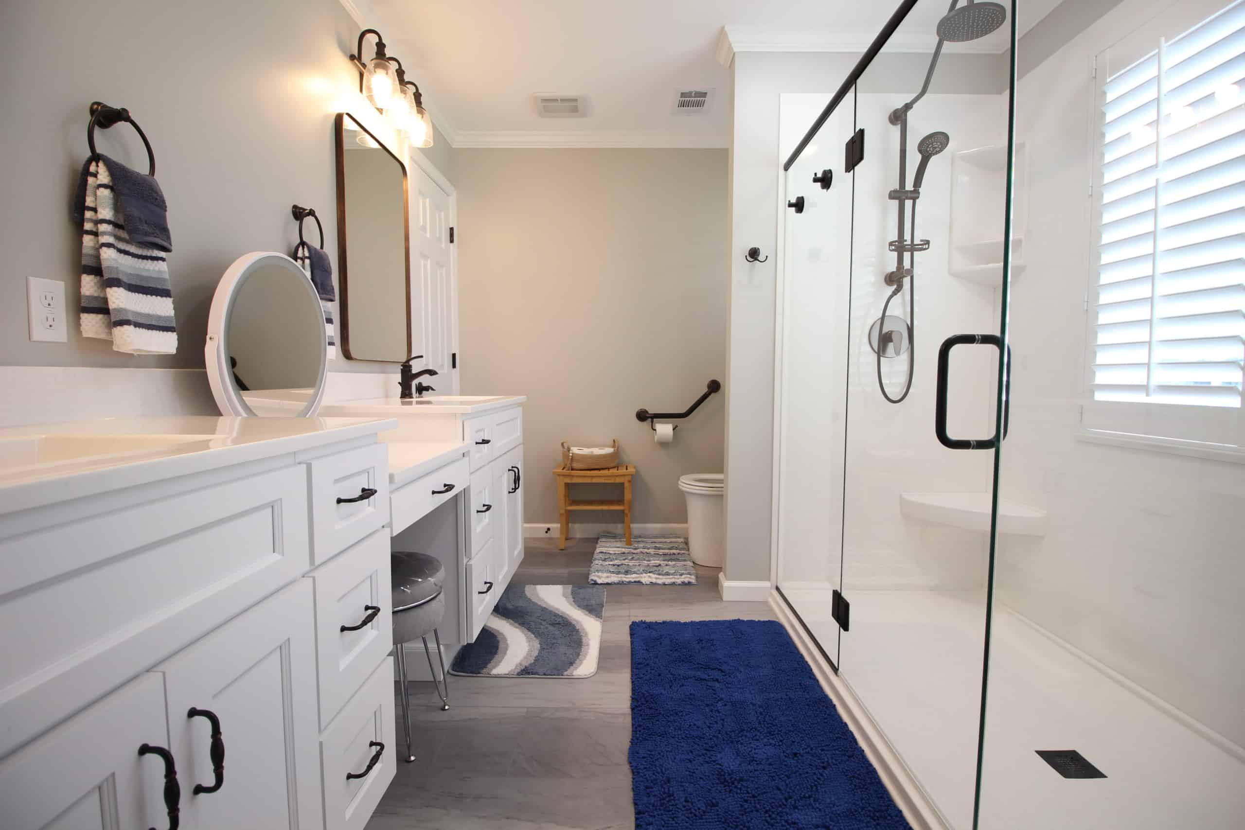 Bright bathroom featuring double sinks, mirrors, a glass shower, blue and white rugs, towel racks, and a separate toilet area. A wooden stool sits near the back wall by a window with blinds.